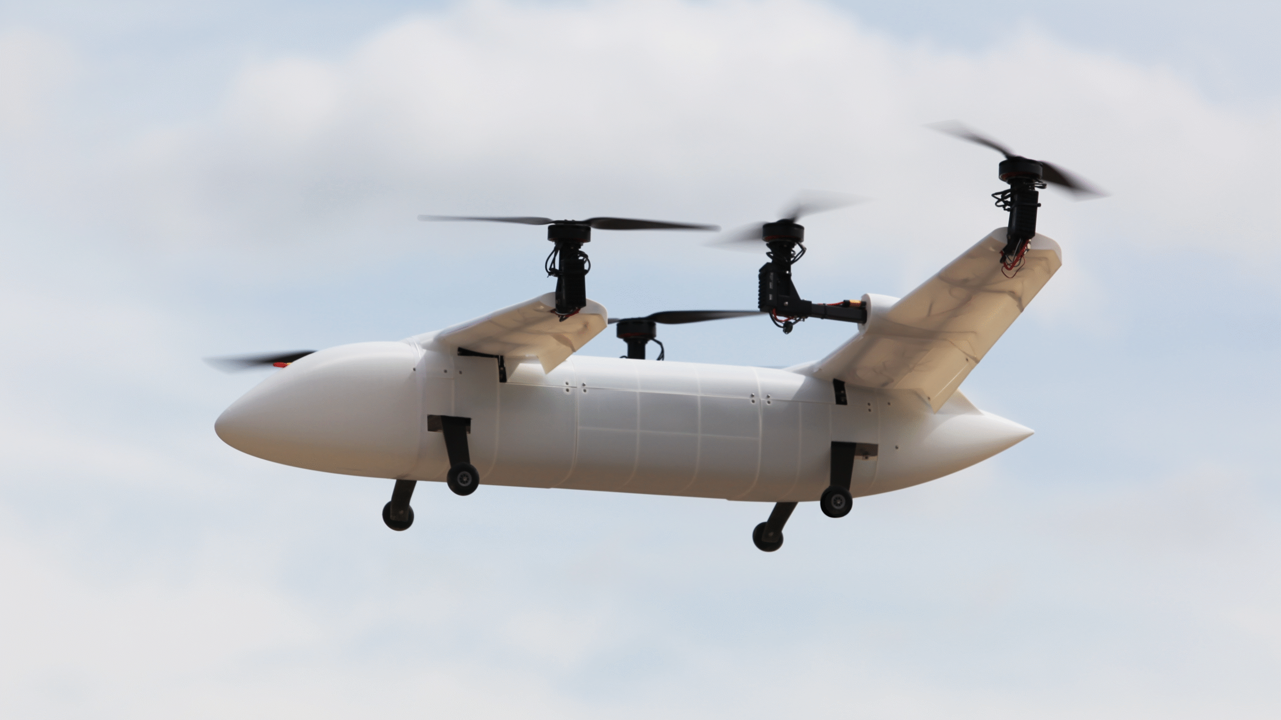 Quadcopter drone in flight with four propellers and a white fuselage against a cloudy sky, landing gear visible below.